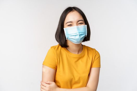 Healthy and happy asian woman in medical face mask, has band aid on shoulder after covid-19 vaccination, got vaccinated from omicron delta variant, whtie background