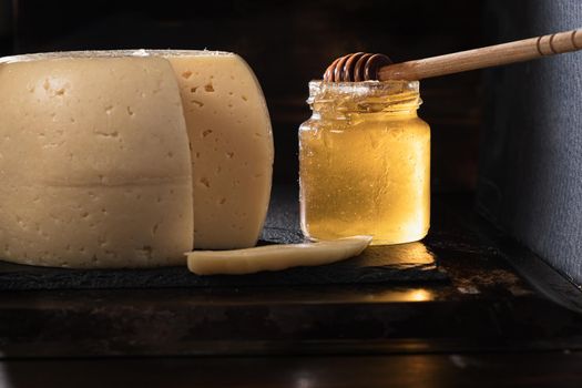 The head of cheese lies on a slate dish lying on a rusty surface. Nearby is a stained jar of honey in which the Honey Stir Bar. A piece of cheese sliced by a pockmark. Horizontal orientation.