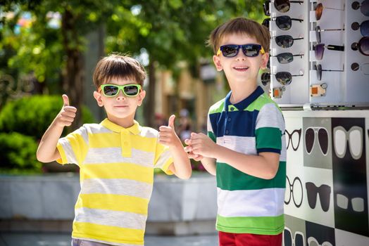 A child and a choice of sunglasses. Two little boys are standing in sun-proof glasses against the background of a shop window with glasses. Kid help choose each other. Sunglasses sale during summer vocation