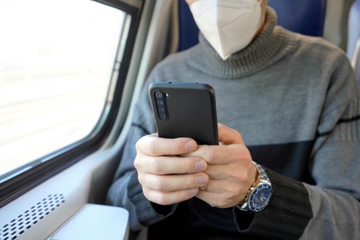 Man on public transport using mobile app wearing medical face mask. Train commuter holding cellphone with mandatory protective mask KN95 FFP2. Focus on the phone.