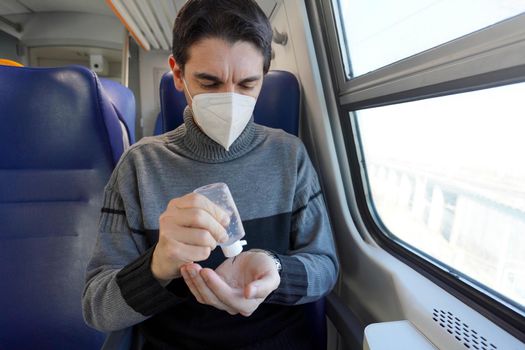 Travel safely on public transport. Young man with protective face mask disinfects hands from alcohol gel dispenser. Passenger with medical mask sanitizing hands inside train carriage. 