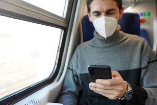 Businessman on public transport using smartphone app wearing protective face mask. Train passenger holding mobile phone during travel commute wearing KN95 FFP2 face mask. Focus on the phone.