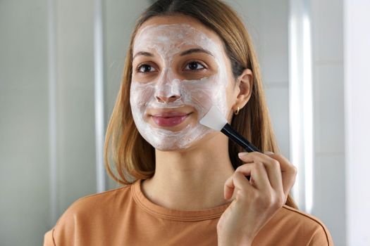 Young woman applying moisturizing mask with facial sheet brush looks herself in the mirror at home