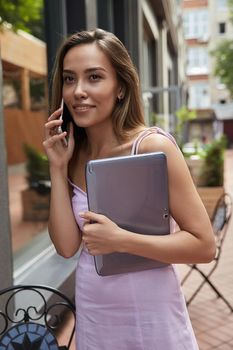 young asian woman in dress standing outdoors with laptop, calling by smartphone