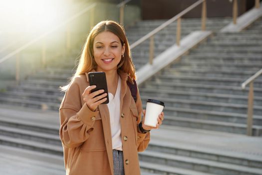 Smiling young business woman holding smartphone and take away coffee in the morning with blurred stairs on background