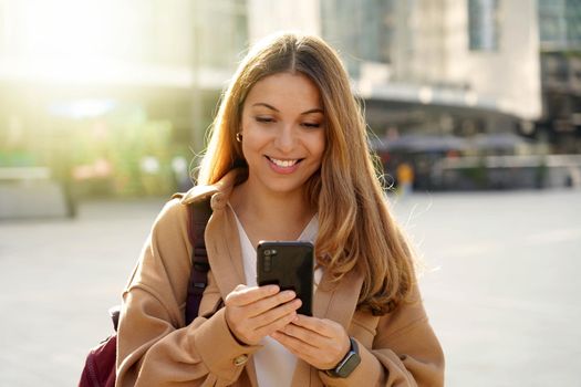 Young cheerful woman using cell phone and texting message on city street. Happy girl holding smart phone in hands and smiling. Surfing internet outdoors. Online communication.