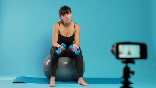 Fitness vlogger recording workout video on camera in studio, using dumbbells to explain lifting technique for sport practice. Muscular woman filming online training lesson on toning ball.
