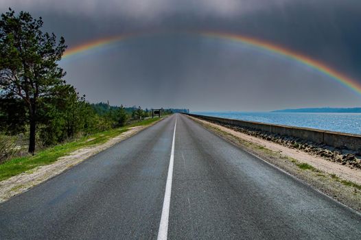 A rainbow with a thundercloud on the horizon of a highway.