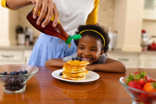 Midsection of mother putting maple syrup on pancake of african american daughter in kitchen at home
