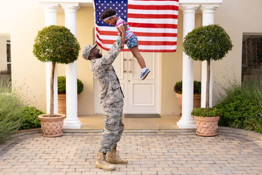 Happy african american military father holding aloft daughter in front of house with usa flag