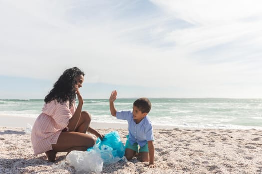 Happy biracial mother and son giving high-five while collecting garbage in bag at beach against sky