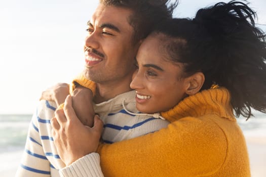 Smiling biracial young woman embracing boyfriend from behind while looking away at beach