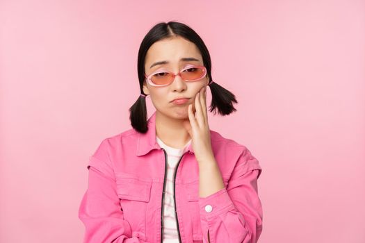 Image of sad asian girl sulking, touching her cheek, pouting disappointed, has toothache, standing over pink background