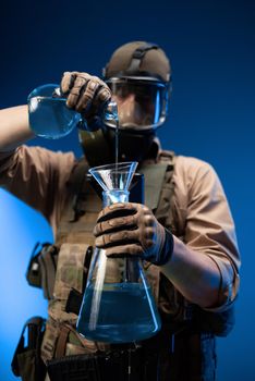a man in military clothes and a gas mask with laboratory chemical flasks pours reagents