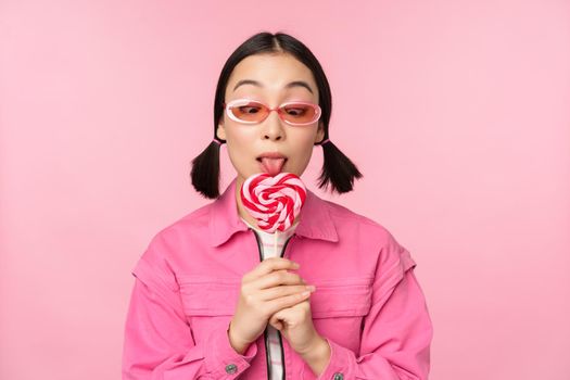 Silly and cute asian female model licking lolipop, eating candy sweet and smiling, looking excited, standing over pink background