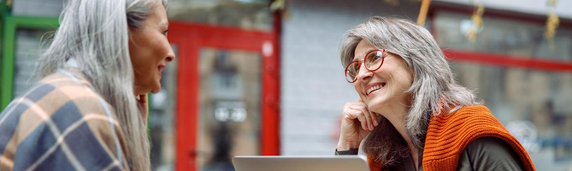 Grey haired lady with laptop spends time with Asian friend on outdoors cafe terrace