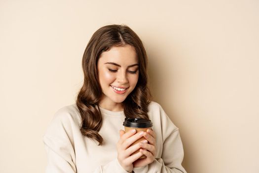 Beautiful authentic woman smiling, holding warm cup of coffee and looking happy, standing over beige background