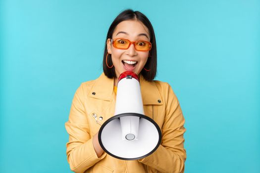 Stylish asian girl making announcement in megaphone, shouting with speakerphone and smiling, inviting people, recruiting, standing over blue background