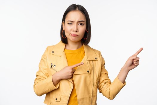 Disappointed asian girl frowning, sulking upset, pointing fingers right at advertisement, standing over white background