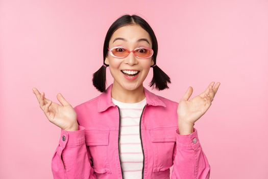 Image of asian girl looking surprised and excited, smiling, amazed reaction to big news, standing over pink background