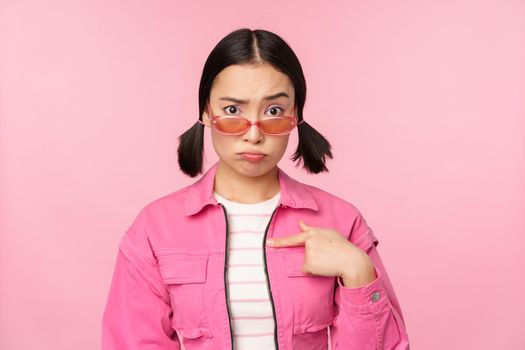 Portrait of asian girl looks confused and points at herself, perplexed face, stares with disbelief at camera, stands over pink background