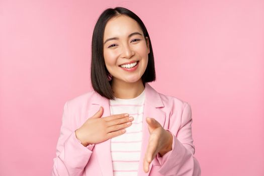 Portrait of smiling, pleasant businesswoman shaking hands with business partner, handshake, extending hand and saying hello, standing over pink background
