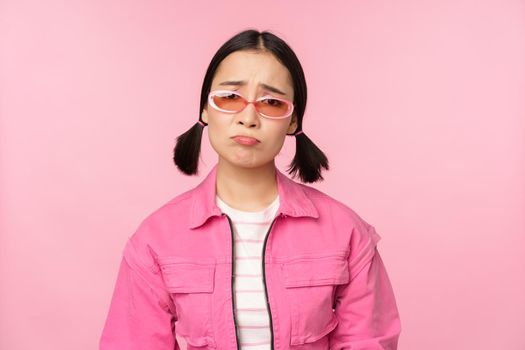 Portrait of sad and gloomy asian girl sulking from disappointment, standing upset against pink studio background