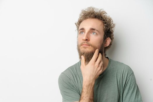 Thoughtful or pensive young handsome bearded wild curly hair man with bright blue eyes isolated on white background. Young thinking man in green tshirt on white