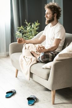 Handsome man sitting on the couch or sofa at home, enjoying meditation in his pajamas in living room during lockdown or being sick or weekend relax