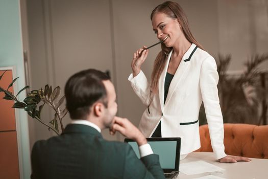Young woman sitting on the table and sexually looking at young man. Two young and attractive colleagues man and woman flirting at office. Flirting in the workplace. Office romance