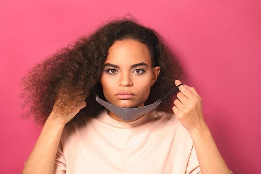No emotion or indifference holding reusable mask African American girl in peachy t-shirt, to prevent others from corona COVID-19 and SARS cov 2 infection isolated on pink background