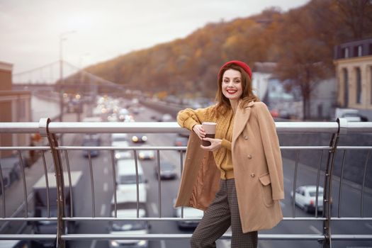 Portrait of a pretty smiling french young woman looking at camera standing at pedestrian bridge with cars on the road. Looking happy fashion girl in a ed beret and beige coat