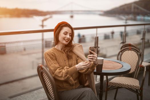 Video call beautiful young woman using her smartphone while having cup of coffee. Frenchwoman wearing red beret sitting on terrace of restaurant or cafe with background of autumn urban city
