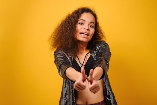 Hands front and thumbs up happy Afro American girl with beautiful hairstyle posing smile looking side away with hand on hips wearing shiny black jacket and black top on yellow background