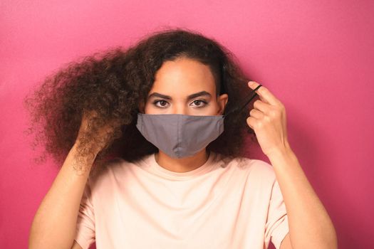 Putting on a reusable face mask young African American female in light peachy t-shirt, to prevent others from corona COVID-19 and SARS cov 2 infection isolated on pink background