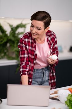Woman in kitchen using laptop holding eggs in hand smiling while reading recipe or having a video call. Woman in red plaid shirt in modern kitchen interior