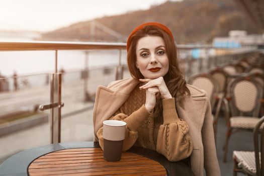 Lovely french young woman sitting at restaurant terrace with coffee mug looking at camera. Portrait of stylish young woman wearing autumn coat and red beret outdoors