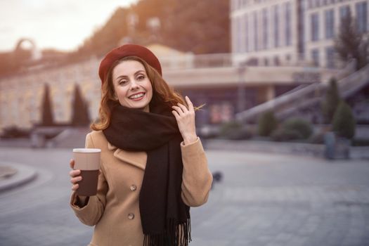 Casual portrait of french woman holding coffee mug outside in autumn beige coat and red beret with urban city background. Tinted photo