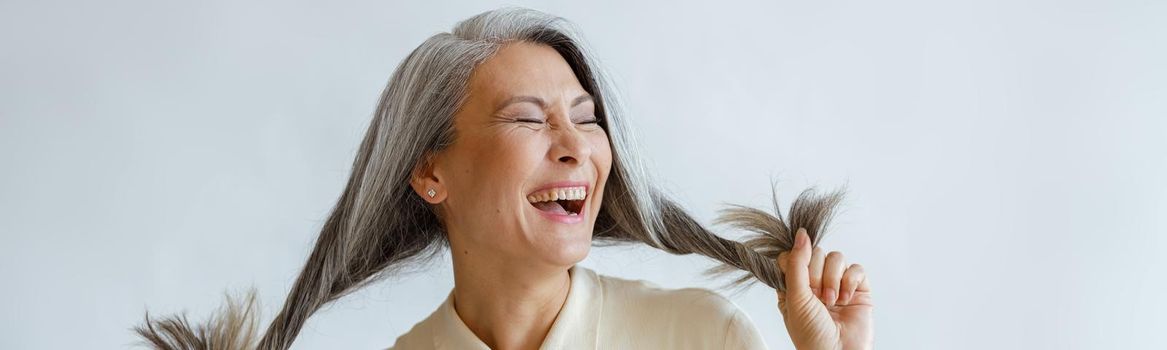 Asian lady holds long hoary hair laughing on light grey background