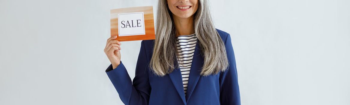 Pretty Asian lady with grey hair holds card with word Sale on light background