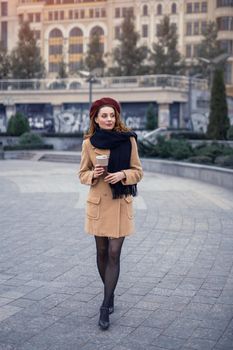 Beautiful french styled young woman with coffee mug on the street female fashion. Portrait of stylish young woman wearing autumn coat and red beret outdoors. Autumn accessories