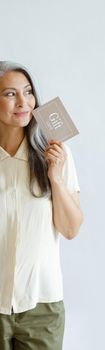 Thoughtful hoary haired Asian lady customer holds gift card standing on light grey background