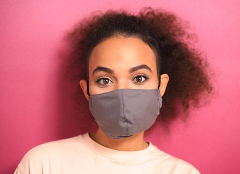 Indifference look African American girl in reusable mask and peachy t-shirt. Stressed or shocked look and prevent others from corona COVID-19 and SARS cov 2 infection isolated on pink