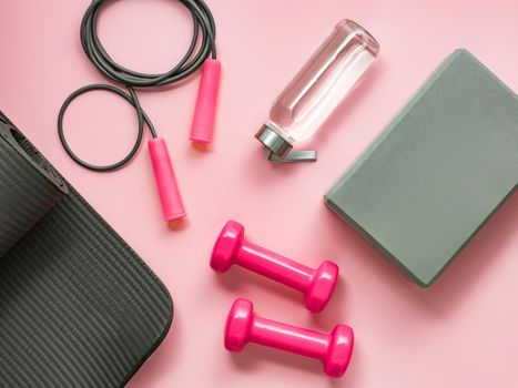 Stylish gray and pink fitness training and gym flat lay. Top view of gray sport mat, yoga block, skipping rope, pink dumbbells, water bottle on pink background. Set for pilates, fitness, yoga