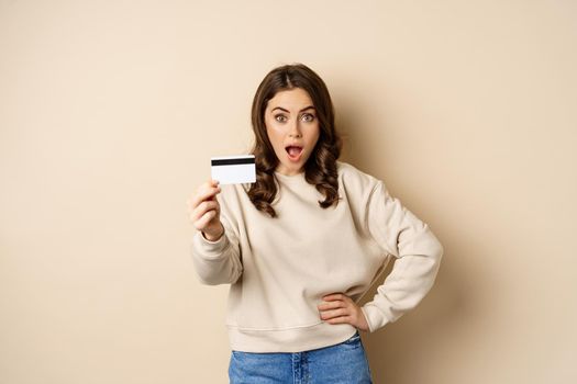 Surprised woman showing credit card, gasping impressed, impression of discounts or shopping, standing over beige background