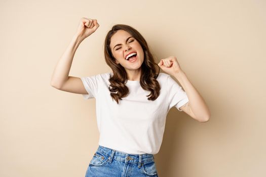 Enthusiastic adn surprised woman winning, dancing and celebrating, standing in tshirt over beige background