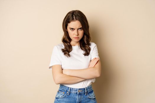Offended angry woman cross hands on chest, frowning and sulking insulted, standing over beige background