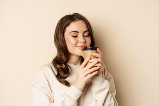 Woman smiling and smelling delicious cup of coffee in takeaway cup, standing over beige background