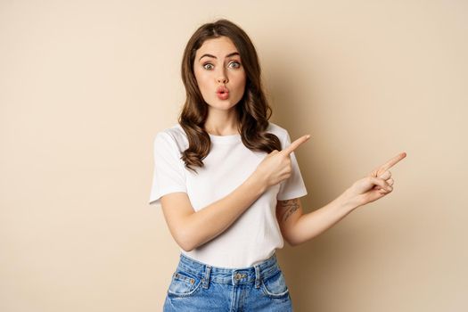 Excited girl pointing fingers right, showing promo with curious face expression, standing over beige background