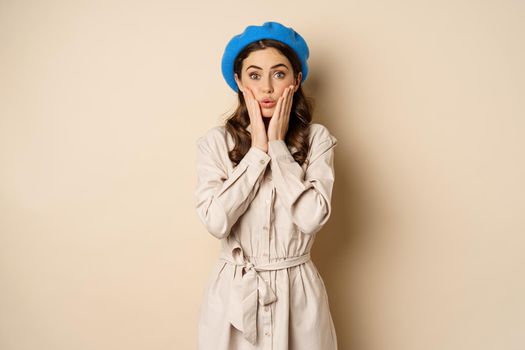 Image of stylsh beautiful woman looking surprised, shocked reaction at camera, posing in trench coat against beige background